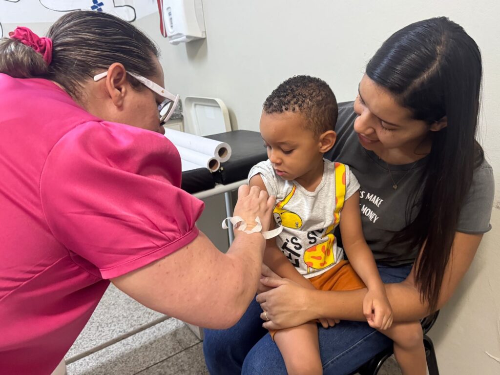 A imagem mostra uma cena em um consultório médico. Uma profissional de saúde, vestida com uma blusa rosa, está aplicando uma injeção no braço de um menino que está sentado no colo de uma mulher. O menino, que veste uma camiseta cinza com estampa de girafa e shorts laranja, olha para o braço com uma expressão apreensiva. A mulher, com cabelos escuros e longos, veste uma camiseta cinza com uma inscrição e olha para o menino com um sorriso reconfortante. Ao fundo, há uma maca com rolos de papel e uma parede branca. A atmosfera geral sugere um momento de cuidado médico para a criança, com o apoio de sua mãe.