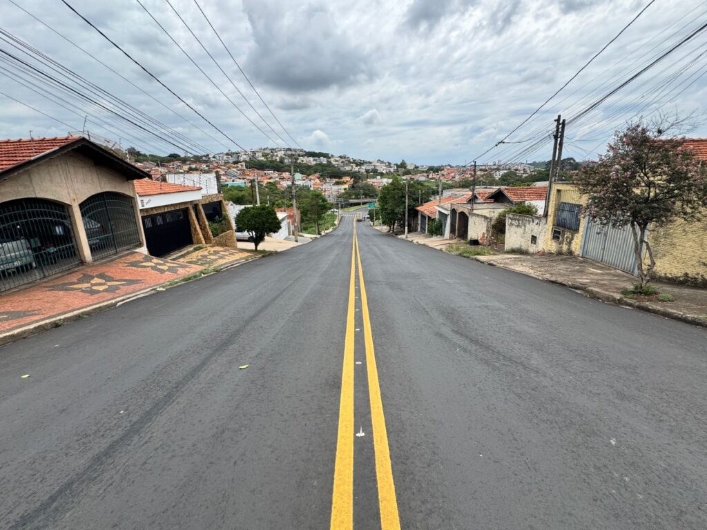 A imagem mostra uma rua asfaltada com duas faixas amarelas pintadas no centro, que se estendem em direção a uma paisagem urbana no topo de uma colina. A rua está ladeada por casas com telhados de barro e muros baixos. A céu está nublado, com nuvens espessas que cobrem a maior parte do céu. Fios elétricos e postes de luz estão visíveis ao longo da rua, criando um padrão em zigue-zague contra o céu. A perspectiva da rua, com o ponto de fuga no horizonte, dá uma sensação de profundidade e distância. As casas nas laterais da rua variam em estilo e condição, com algumas parecendo bem cuidadas e outras mostrando sinais de desgaste. Uma árvore com flores rosadas está à direita, adicionando um toque de cor à cena.