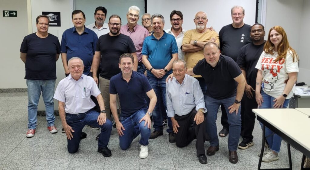 A imagem mostra um grupo de 17 pessoas, sendo 16 homens e 1 mulher, posando para uma foto em um ambiente interno. Algumas estão de pé na parte de trás, enquanto outras estão agachadas na frente. O local parece ser um escritório ou sala de reunião, com um sinal de Wi-Fi e um sinal de saída visíveis ao fundo.