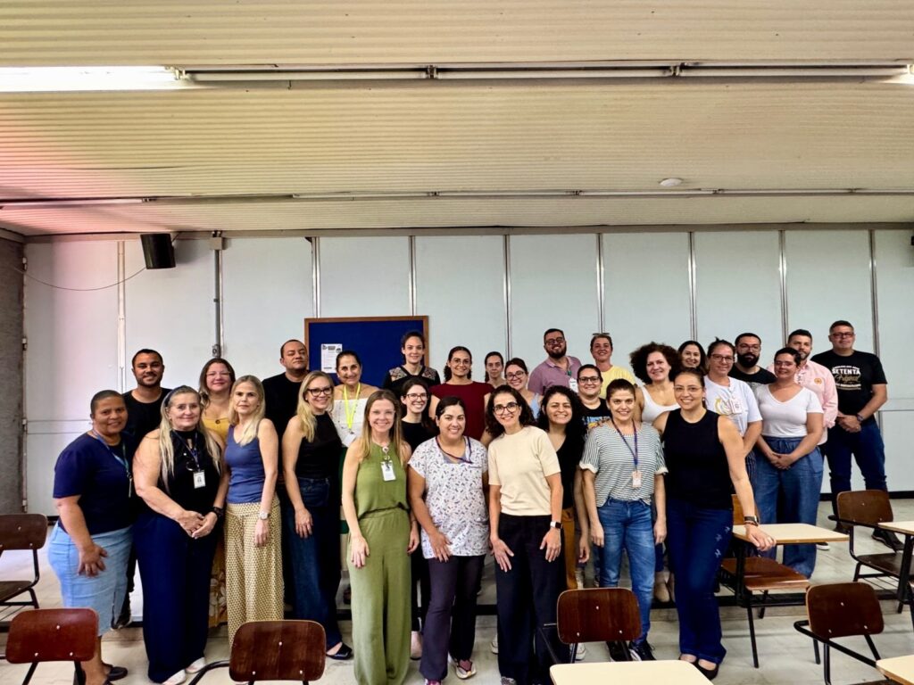 A imagem mostra uma sala de aula com um grupo de pessoas sentadas em carteiras, aparentemente participando de uma palestra ou aula. Duas mulheres estão em pé na frente da sala, uma delas falando e gesticulando, enquanto a outra observa. O ambiente parece ser uma universidade ou centro de treinamento, com as carteiras dispostas em degraus e uma grande janela com cortinas ao fundo. A iluminação é proveniente de lâmpadas no teto, e há um projetor pendurado. As pessoas na plateia têm idades variadas e vestem roupas casuais. Algumas parecem atentas à apresentação, enquanto outras conversam ou olham para o lado. Há bolsas e cadernos sobre as carteiras.