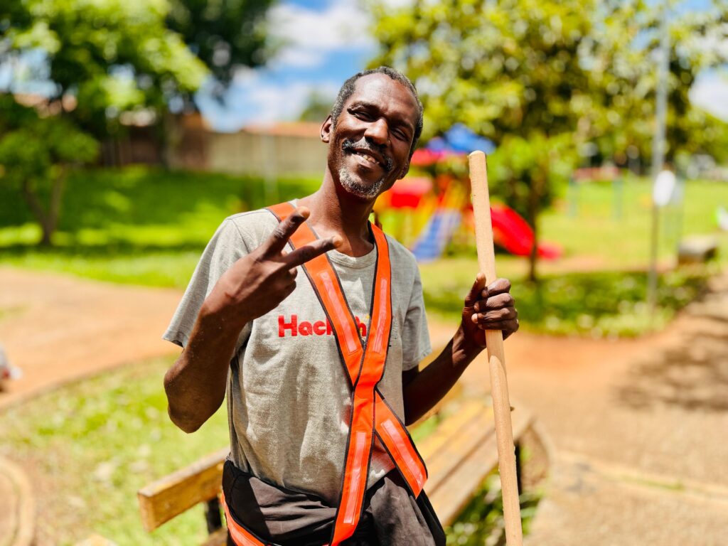 Um homem com pele escura, barba e bigode grisalhos, sorri para a câmera, fazendo um sinal de paz com a mão esquerda. Ele veste uma camiseta cinza com um colete laranja de alta visibilidade por cima. O colete tem detalhes refletivos e a palavra "Hack" escrita em vermelho no peito. Ele segura um pedaço de madeira em sua mão direita. O fundo da imagem mostra um parque ou área recreativa com grama verde, árvores e um parquinho infantil com escorregadores vermelhos e azuis. O céu está parcialmente nublado. A imagem é tirada em um dia ensolarado, com a luz do sol projetando sombras e realçando as cores.