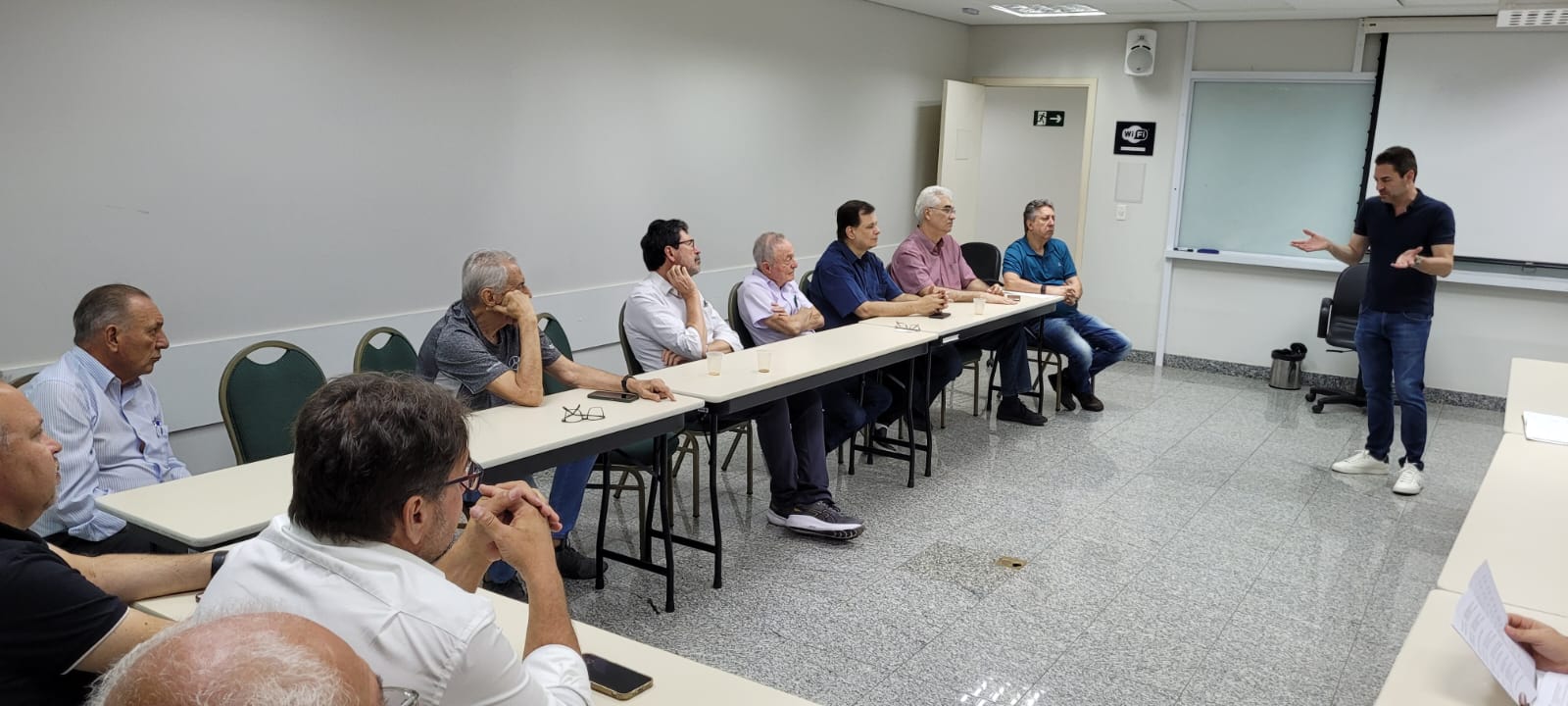 Um homem está de pé em uma sala de reuniões, gesticulando com as mãos como se estivesse explicando algo para um grupo de homens sentados à mesa.