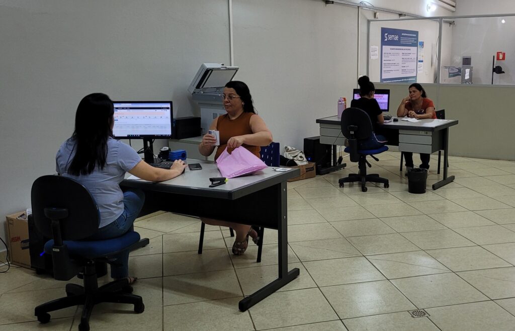A imagem mostra um escritório com quatro pessoas em três mesas diferentes, todas sentadas em cadeiras de escritório giratórias. Na mesa da frente, uma mulher com cabelo preto comprido está sentada de costas para a câmera, olhando para um monitor de computador. Ela usa uma camisa azul clara e jeans. Ao lado dela, outra mulher, vestida com uma blusa marrom sem mangas, está sentada de frente para a câmera. Ela está segurando algumas cartas ou cartões na mão direita e um saco rosa na mão esquerda, parecendo estar em meio a uma transação ou entrega. Uma impressora está posicionada atrás das duas mulheres. Mais atrás, em outra mesa, uma mulher com cabelo preto preso em um coque está sentada de costas para a câmera, olhando para um monitor de computador. Ela usa uma camisa preta e jeans. Ao lado dela, sentada de frente para a câmera, está uma mulher com cabelo preto, vestida com uma blusa laranja e calça preta. Ela está apoiada na mão e parece estar conversando com a pessoa à sua frente. Um grande cartaz com o logotipo "SEMAE" está fixado na parede de vidro ao fundo, indicando que este pode ser um escritório de atendimento ao cliente ou administrativo. O chão do escritório é revestido com ladrilhos de cor creme e há uma grade de ventilação no chão. A iluminação é artificial, vindo de luminárias no teto.