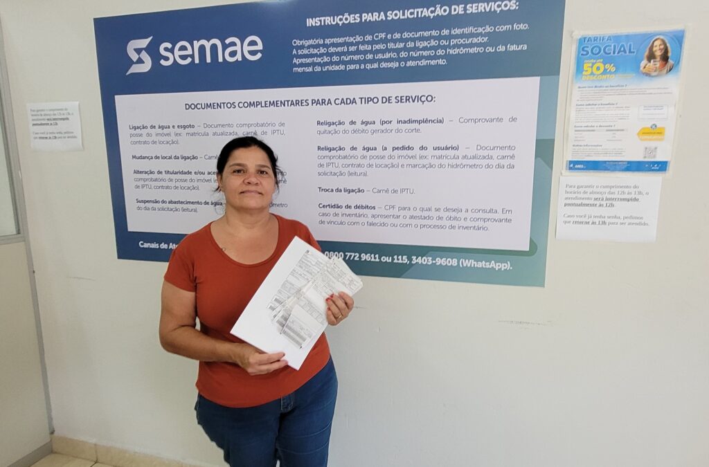A mulher, vestida com uma blusa laranja e calça jeans, está em pé em frente a um grande painel informativo com o logotipo da SEMAE. Ela segura alguns papéis nas mãos. O painel contém instruções para solicitação de serviços, documentos complementares para cada tipo de serviço e informações sobre a Tarifa Social com 50% de desconto. Há também um aviso sobre o horário de almoço, indicando que o atendimento é interrompido pontualmente às 12h e retorna às 13h. Números de telefone e WhatsApp para contato estão exibidos na parte inferior do painel. O ambiente parece ser um escritório ou balcão de atendimento público.
