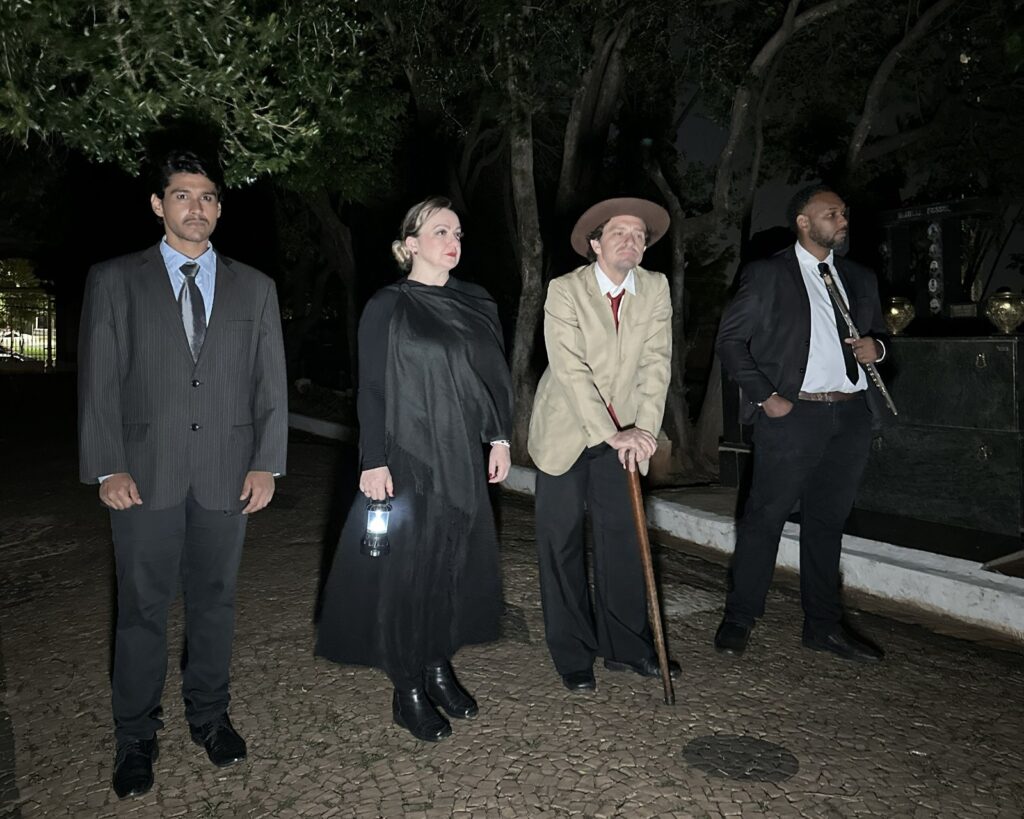 A cena noturna retrata quatro pessoas em um ambiente que parece ser um cemitério ou um local com túmulos. A iluminação é escassa, vindo principalmente de uma lanterna que uma das mulheres segura. À esquerda, um homem vestido com terno risca de giz cinza e camisa azul clara com gravata escura está em pé, olhando para frente. Ao seu lado, uma mulher com um longo vestido preto e um xale cobrindo seus ombros, segura uma lanterna acesa na mão direita. No centro, um homem com chapéu marrom, blazer bege, camisa branca e gravata vermelha apoia-se em uma bengala de madeira, com uma expressão pensativa. À direita, outro homem veste um terno preto, camisa branca e gravata, segurando o que parece ser uma flauta ou um instrumento similar. Ele também olha para a frente, com as mãos nos bolsos. Ao fundo, vislumbra-se a estrutura de alguns túmulos ou monumentos funerários, com urnas decorativas e inscrições. As árvores com folhagem escura criam um pano de fundo sombrio para a cena. O chão é pavimentado com pedras irregulares.
