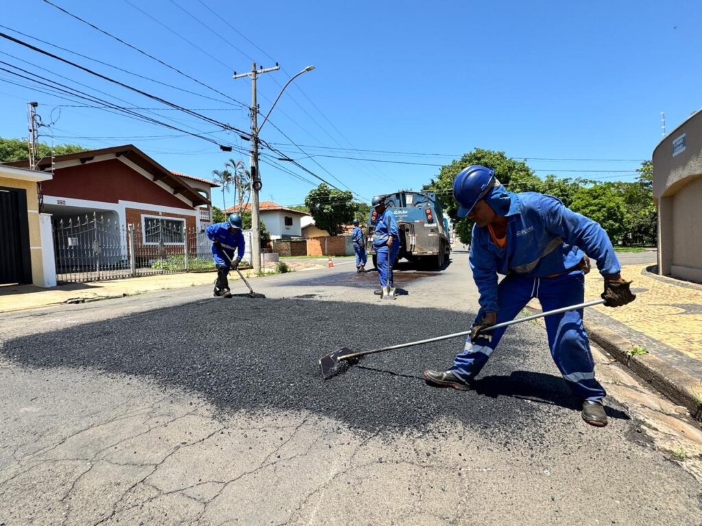 A imagem mostra uma equipe de trabalhadores em um dia ensolarado, realizando reparos em uma rua de asfalto. No primeiro plano, um trabalhador vestido com uniforme azul e capacete azul está espalhando asfalto quente com uma ferramenta longa. Ele usa luvas e botas de segurança. Mais atrás, outros dois trabalhadores estão envolvidos no mesmo trabalho. Um deles também usa uniforme azul e capacete, enquanto o outro, mais ao fundo, usa um colete laranja sobre o uniforme. Uma caminhonete de asfalto está estacionada na rua, com sua traseira aberta, indicando que está descarregando o material. O cenário é uma rua residencial com casas de um lado e árvores do outro. O céu está azul claro e o sol brilha intensamente, projetando sombras nítidas no chão. A calçada ao lado da rua tem um padrão de mosaico. A cena geral transmite a ideia de trabalho pesado e a manutenção da infraestrutura urbana.