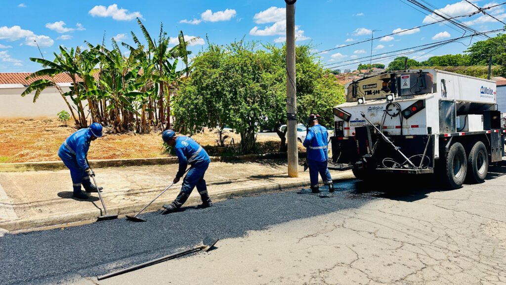 A imagem mostra uma equipe em ação realizando obras de pavimentação asfáltica em uma rua residencial. Dois trabalhadores, vestidos com uniformes azuis e capacetes, utilizam ferramentas para espalhar e nivelar o asfalto recém-aplicado. Um caminhão especializado em pavimentação, com a marca "Molise", está posicionado ao lado da área de trabalho, fornecendo o material. Ao fundo, observam-se casas com telhados vermelhos, árvores frondosas e um céu azul com nuvens brancas, indicando um dia ensolarado. A cena transmite a ideia de trabalho em equipe e a execução de serviços de infraestrutura urbana.