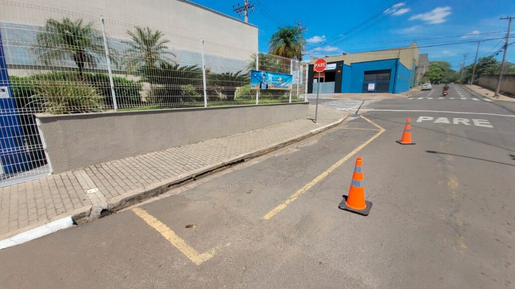 A imagem mostra uma rua em um dia ensolarado, com um céu azul claro e algumas nuvens. * No lado esquerdo, há um prédio com uma parede cinza e uma cerca de metal. Plantas e palmeiras crescem atrás da cerca. Há também um outdoor com a imagem de um homem e texto em português, anunciando uma escola. * No lado direito da rua, há um prédio azul com uma entrada preta. Um sinal de "PARE" está visível em vermelho. * A rua tem marcações amarelas e brancas no asfalto. Há duas laranjas cones de trânsito na estrada, um mais perto da câmera e outro mais longe. * Ao fundo, a rua continua com mais prédios e árvores, e alguns veículos são visíveis, incluindo uma motocicleta e um carro. Um sinal de "PARE" também está pintado no asfalto da estrada mais adiante. * O pavimento da calçada é feito de blocos de concreto interligados. Um ralo está visível na calçada, perto da rua. * A imagem parece ter sido tirada de um ponto de vista baixo, olhando para a rua e os prédios.