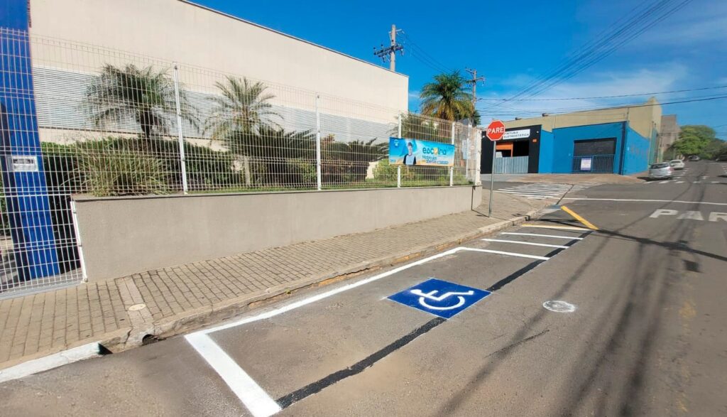 A imagem mostra uma rua em um dia ensolarado, com um céu azul claro e algumas nuvens. Há uma vaga de estacionamento pintada na rua, com o símbolo internacional de acessibilidade para pessoas com deficiência. A vaga é delimitada por linhas brancas e pretas. Ao fundo, há um prédio com uma fachada clara e uma cerca de metal. Há também algumas palmeiras e arbustos. Em um dos prédios, há uma placa com a imagem de um homem e a palavra "Escolha". Mais adiante, há um prédio azul com uma porta de garagem preta. A rua tem uma faixa de pedestres e uma placa de "PARE". Há alguns carros estacionados e em movimento na rua. A imagem parece ter sido tirada de um ângulo baixo, o que dá uma perspectiva interessante da cena.