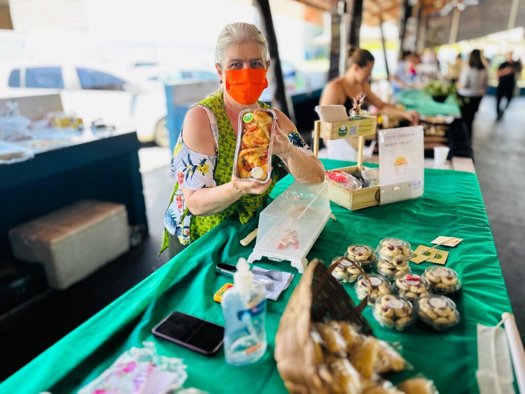 A imagem mostra uma mulher usando uma máscara laranja e um colete verde vibrante sobre uma blusa estampada. Ela está em uma barraca, segurando uma embalagem transparente com comida, que parece ser pão de queijo ou algo semelhante. A mesa à frente dela está coberta com uma toalha verde brilhante e exibe vários outros produtos alimentícios, incluindo biscoitos em embalagens plásticas e um cesto com pães. Há também um frasco de desinfetante para as mãos, um celular e um pequeno cartaz com uma imagem de sorvete e o preço "R$ 12,00". Ao fundo, outras pessoas estão em barracas, sugerindo um mercado ou feira.