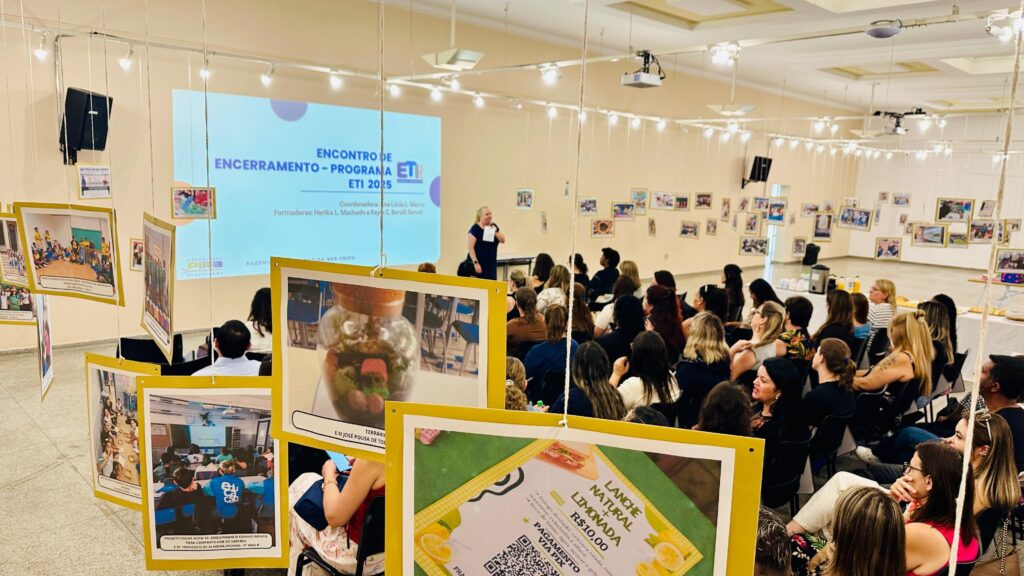 A imagem mostra um evento de encerramento de um programa chamado ETI 2025. Há uma apresentação em um telão ao fundo, onde uma mulher está falando para uma plateia sentada em cadeiras. Fotos e cartazes estão pendurados em fios, decorando o ambiente. Um dos cartazes em destaque exibe um "LANCHE + NATURAL LIMONADA" por R$10,00, com opções de pagamento via PIX. Há também um terrário em um dos cartazes. O evento parece ser um encontro de formação ou de partilha de saberes.