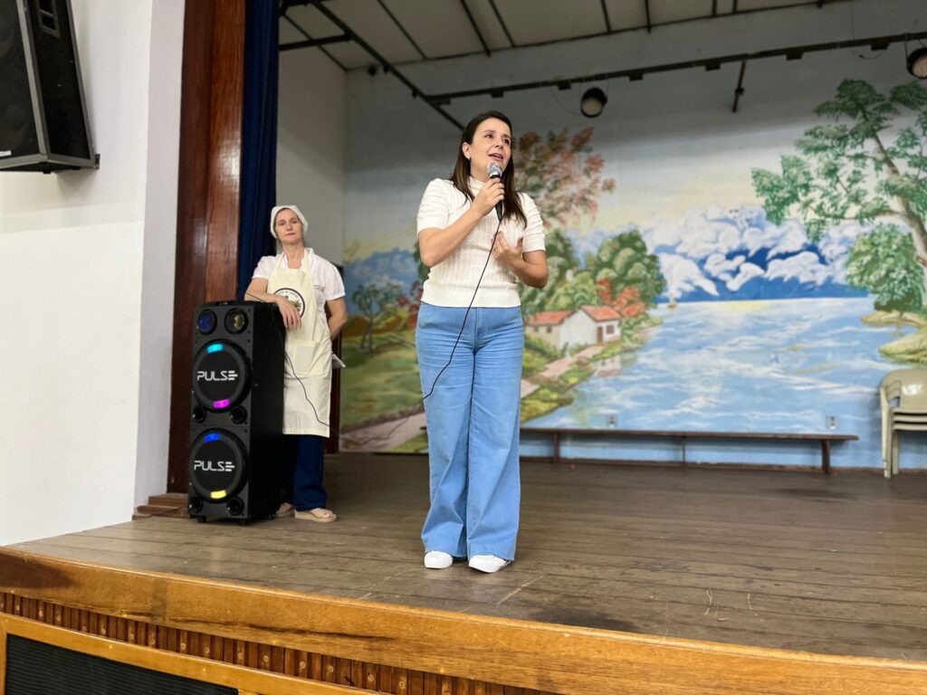 A imagem mostra um palco com duas mulheres. Uma mulher, à direita, está de pé e falando em um microfone. Ela veste uma blusa branca de manga curta e calças azuis largas. A outra mulher, à esquerda, está parada perto de uma caixa de som, usando um avental branco sobre uma roupa escura e um lenço na cabeça. O fundo do palco é decorado com uma pintura colorida que retrata uma paisagem com casas, árvores e um lago ou rio, com montanhas ao fundo. Há uma caixa de som preta com luzes coloridas ao lado da mulher à esquerda. O palco é feito de madeira, com uma estrutura de madeira na frente. Há também uma bancada de madeira e algumas cadeiras ao fundo.