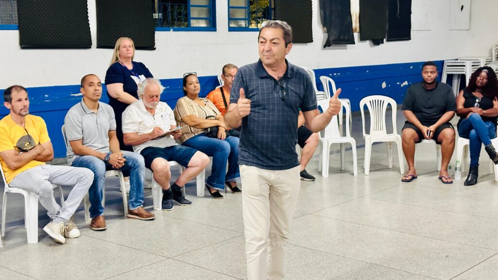 A imagem mostra um grupo de pessoas sentadas em cadeiras brancas, em uma sala com paredes azuis e pretas. No centro, um homem com camisa xadrez e calças claras está em pé, sorrindo e fazendo um sinal de positivo com os dois polegares. Ele parece estar discursando ou apresentando algo para o grupo. À sua esquerda, sentados em cadeiras, estão um homem com camiseta amarela e braços cruzados, outro com camiseta cinza e mãos juntas no colo, e um homem mais velho com camisa branca e barba grisalha, que está escrevendo em uma prancheta. Atrás dele, uma mulher loira com camiseta azul escura observa. À direita do homem em pé, sentados em cadeiras, estão um homem negro com camiseta preta e chinelos, e uma mulher negra com blusa preta e jeans, que olha para o lado. Outras pessoas estão visíveis ao fundo, mas com menos destaque. O ambiente parece ser um local de reunião ou evento comunitário.