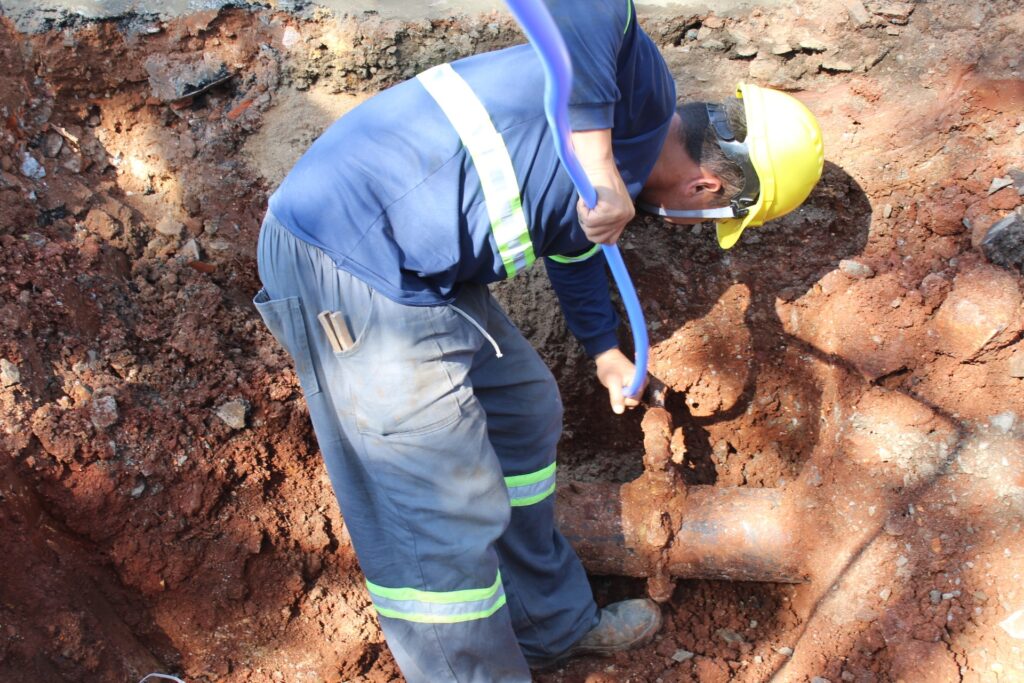 Um trabalhador, vestindo um capacete amarelo e uniforme azul com faixas refletivas, está agachado em uma vala cavada no solo. Ele segura um tubo azul e parece estar trabalhando em uma tubulação de metal enferrujada e antiga que emerge da terra. A vala revela camadas de solo argiloso e pedras, indicando um trabalho de escavação em andamento. Ferramentas parecem estar presas ao bolso da calça do trabalhador.
