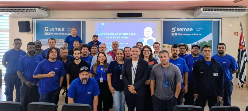 A imagem mostra um grupo de homens e duas mulheres posando para uma foto em um ambiente interno. Todos parecem estar vestindo uniformes azuis, com exceção de alguns indivíduos que usam jaquetas ou blusas pretas e cinzas. Ao fundo, há um banner com o logotipo "semae" e a frase "UM NOVO TEMPO", além de um slogan sobre "A IMPORTÂNCIA DO NOVEMBRO AZUL NA SAÚDE DO HOMEM". Há também uma bandeira do estado de São Paulo à direita. As pessoas estão sorrindo e parecem estar participando de um evento ou reunião.
