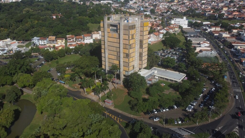 A imagem aérea revela uma paisagem urbana intercalada com áreas verdes exuberantes. No centro, destaca-se um edifício alto e moderno, com uma fachada composta por elementos verticais em tons de bege e amarelo, que se ergue em meio a uma vegetação densa. Ao redor do edifício, um estacionamento com vários carros e um gramado bem cuidado. À esquerda, um lago de formato irregular reflete o verde das árvores que o circundam. Mais adiante, um conjunto de casas com telhados vermelhos e brancos compõe um bairro residencial, com ruas e mais vegetação espalhadas. À direita, a cidade se estende com mais casas e edifícios, além de uma rua movimentada com carros transitando. Um campo de futebol e outros estacionamentos também são visíveis, integrando a área urbana à natureza. A imagem transmite uma sensação de harmonia entre o construído e o natural.