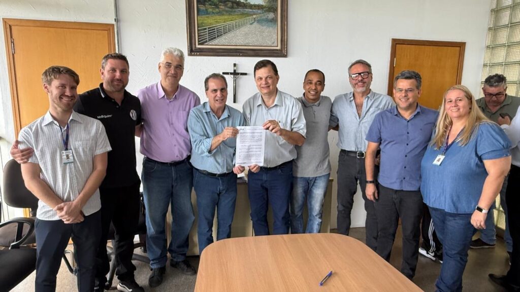 Na imagem, um grupo de dez pessoas, composto por oito homens e uma mulher, posa para uma foto em um escritório. A maioria dos homens está vestida com camisas sociais e calças jeans, enquanto as mulheres usam blusas. No centro da imagem, dois homens seguram um documento, possivelmente um contrato ou acordo, com um sorriso. Ao fundo, há uma porta de madeira, uma pintura na parede e uma cruz religiosa. A iluminação é natural, vinda de uma janela no canto superior direito, e a atmosfera geral parece ser de formalidade e satisfação.