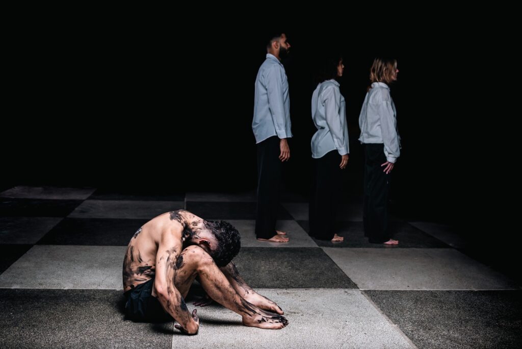 A imagem mostra uma cena dramática e carregada de emoção. Em primeiro plano, há um homem nu da cintura para cima, agachado no chão, coberto de lama escura que cobre seu corpo e rosto. Ele está em uma posição curvada, com a cabeça baixa, transmitindo uma sensação de angústia ou sofrimento profundo. O chão onde ele está é um tabuleiro quadriculado em tons de cinza claro e escuro, com uma textura que lembra mármore ou pedra. Ao fundo, em contraste, estão três pessoas — duas mulheres e um homem — todas vestidas com camisas brancas e calças pretas, olhando para o lado oposto do homem no chão. Eles estão em pé, alinhados e com posturas eretas, parecendo indiferentes ou distantes da situação do homem à frente. A iluminação é dramática, com um foco forte sobre o homem no chão, destacando sua figura e a lama, enquanto o fundo está mais escuro, criando uma sensação de isolamento e separação entre ele e as outras pessoas. A composição da imagem sugere temas como sofrimento, solidão, alienação e a distância entre a luta individual e a indiferença social. A cena é intensa e provoca reflexão sobre a condição humana e o isolamento emocional.