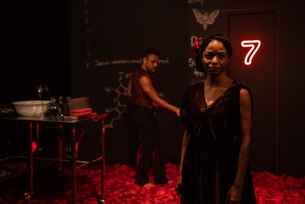A imagem retrata uma cena teatral com iluminação dramática em tons de vermelho. Em primeiro plano, uma mulher com um vestido escuro e brilhante, adornado com uma flor vermelha no cabelo, olha diretamente para a câmera. Ao fundo, um homem com uma camisa sem mangas e calças escuras está em movimento, de costas para a câmera. O cenário é escuro, com uma parede ao fundo que parece ser um quadro-negro, coberta de anotações e diagramas químicos, incluindo a imagem de uma mariposa. À esquerda, uma mesa de metal com rodinhas exibe um recipiente branco, garrafas e um suporte com tubos de ensaio. Um número "7" em neon vermelho se destaca na parede à direita, próximo à mulher. O chão está coberto por um material vermelho que pode ser tecido ou pétalas, intensificando a atmosfera teatral e misteriosa da cena.