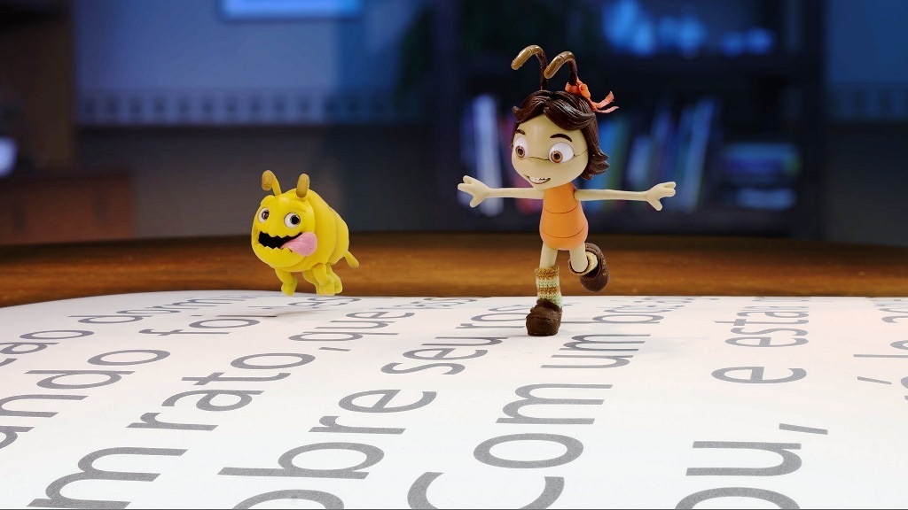 A imagem mostra dois personagens animados sobre uma grande folha de papel com texto impresso. À direita, há uma menina com cabelo castanho preso em um rabo de cavalo com uma fita vermelha. Ela usa um vestido laranja, meias listradas e sapatos marrons, com um sorriso largo e olhos grandes, aparentando estar em movimento, como se estivesse correndo ou dançando. Ao lado dela, à esquerda, está uma pequena criatura amarela, parecida com uma lagarta ou larva, com olhos grandes, antenas e a língua para fora, também parecendo se mover. O fundo é desfocado, sugerindo um ambiente interno, possivelmente uma sala com móveis e estantes de livros. A folha de papel onde eles estão é coberta por texto em português, impresso em fonte grande e cinza, que parece ser parte de um livro ou documento. A iluminação é suave e quente, destacando os personagens e o texto. A perspectiva da imagem é baixa, como se o observador estivesse no mesmo nível dos personagens sobre o papel, criando uma atmosfera lúdica e de história em quadrinhos.