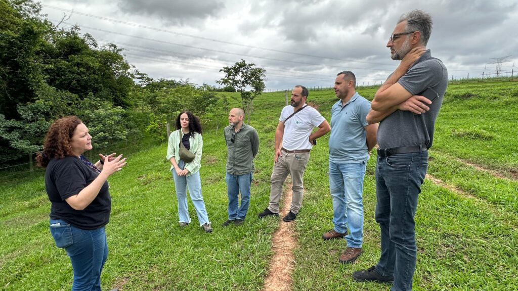 Na imagem, sete adultos estão reunidos em um campo gramado, com árvores ao fundo e um céu nublado acima. À esquerda, uma pessoa gesticula enquanto fala, parecendo conduzir uma explicação ou conversa com os demais, que escutam com atenção. Todos estão vestidos de forma casual, sugerindo um encontro informal ao ar livre. O cenário é rural, com uma trilha de terra visível, reforçando a ideia de que se trata de uma atividade prática em campo.