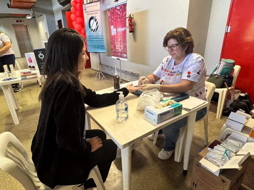 A imagem mostra um evento de testagem de doenças infecciosas, possivelmente em um centro de saúde ou evento comunitário. Duas mulheres estão sentadas em uma mesa. Uma delas, vestida com uma camiseta branca com estampas coloridas, usa luvas e parece estar realizando um teste rápido na outra mulher, que está de blusa preta. Há materiais médicos sobre a mesa, como luvas, algodão e um frasco de álcool em gel. Ao fundo, há cartazes informativos e balões vermelhos, sugerindo um ambiente de campanha de conscientização. Em uma caixa ao lado, pode-se ler "DISTRIBUIÇÃO GRATUITA VENDA PROIBIDA", indicando que os testes ou materiais são oferecidos gratuitamente.