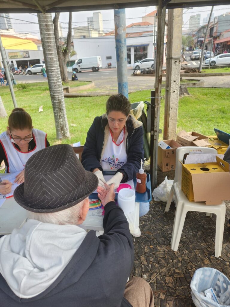A imagem mostra um evento de teste rápido de HIV ao ar livre, possivelmente em uma praça ou rua. Duas mulheres, vestindo aventais brancos com o logo "do HIV F este", estão sentadas atrás de uma mesa, realizando testes em pessoas. Uma delas está com luvas e parece estar coletando uma amostra de sangue do dedo de um idoso, que usa um chapéu. A outra mulher, à esquerda, está anotando informações em uma prancheta. Na mesa, há materiais como recipientes para descarte, um frasco com líquido (possivelmente álcool ou antisséptico) e caixas de papelão com suprimentos. Ao fundo, vê-se uma rua com carros, edifícios e algumas árvores, indicando um ambiente urbano. O clima parece ser diurno e nublado. O evento parece ser uma iniciativa de saúde pública para conscientização e testagem do HIV.