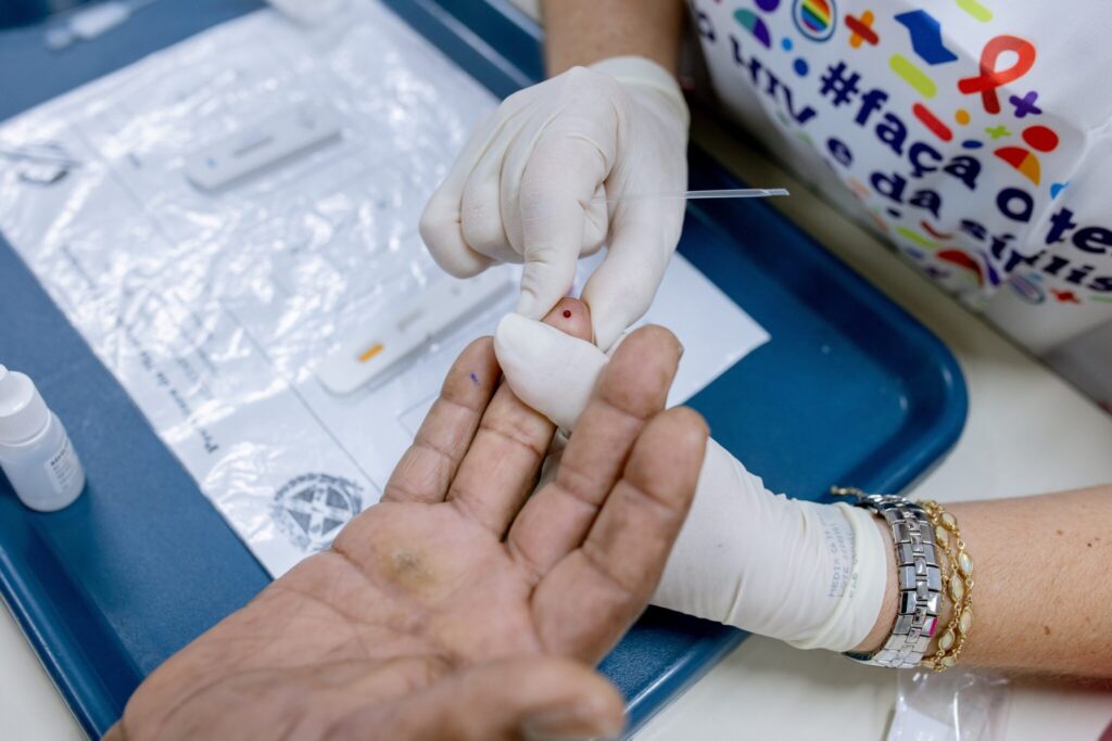 A imagem mostra um close-up de um profissional de saúde, usando luvas brancas, coletando uma gota de sangue do dedo de um paciente. A gota de sangue é visível na ponta do dedo do paciente, e o profissional de saúde está usando um pequeno aplicador para coletar o sangue. No plano de fundo, há um kit de teste rápido para diagnóstico, provavelmente para HIV ou outra doença infecciosa, e um frasco de reagente. A enfermeira usa uma camiseta com um padrão colorido, incluindo um laço vermelho, que pode indicar uma campanha de conscientização sobre saúde. O ambiente parece ser uma clínica ou centro de saúde, com uma bandeja azul contendo os materiais médicos. O paciente tem a mão apoiada, e uma marca de tinta azul é visível em seu dedo.