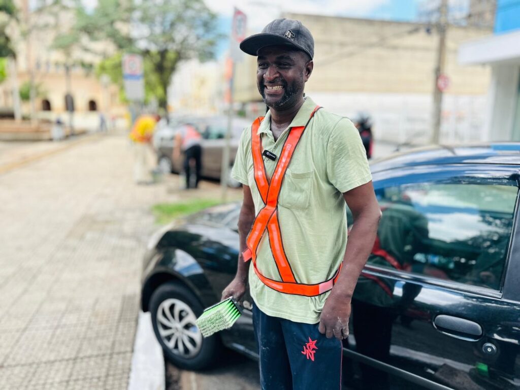 A imagem mostra um homem sorrindo, vestindo uma camisa polo verde clara, um colete laranja de alta visibilidade e um boné escuro. Ele está em pé ao lado de um carro preto, segurando uma escova verde em sua mão direita. O fundo sugere uma rua urbana com outros carros e edifícios. O homem parece ser um trabalhador, possivelmente um limpador de carros ou um trabalhador de rua, dado o colete de alta visibilidade e a escova. A iluminação indica que é dia, com algumas nuvens no céu.