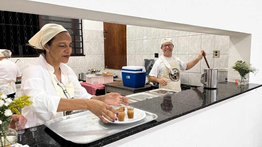 A imagem mostra duas pessoas em uma cozinha, ambas usando toucas e aventais, sugerindo que estão envolvidas na preparação de alimentos. Na frente, uma mulher com um lenço na cabeça e um avental claro sobre uma camisa branca está organizando uma bandeja com quatro pequenos copos contendo um líquido marrom, possivelmente uma bebida ou sobremesa. Seus óculos estão pendurados no avental. Ao fundo, um homem sorridente, também de touca e avental, mexe um grande recipiente metálico com uma colher de pau. Ele usa óculos e parece estar em meio a alguma tarefa culinária. A cozinha parece ser um espaço funcional, com bancadas de granito, azulejos nas paredes e vários utensílios e recipientes visíveis, incluindo uma caixa térmica azul e recipientes brancos. Há também um pequeno arranjo de flores em um vaso sobre a bancada.