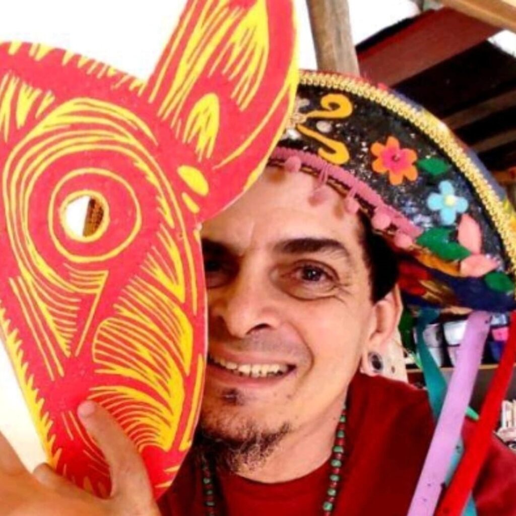 A imagem mostra um close-up do rosto de um homem sorrindo, segurando uma máscara vermelha e amarela na frente do seu rosto. Ele está usando um chapéu colorido e decorado, com pompons e flores. O homem tem barba curta e usa um colar de contas. A máscara que ele segura tem um design abstrato com espirais e um buraco no centro, possivelmente para os olhos. O fundo da imagem está desfocado, mas sugere um ambiente externo ou rústico.