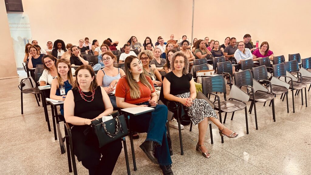 A imagem mostra uma sala de aula ou auditório com um grupo de pessoas, predominantemente mulheres, sentadas em cadeiras voltadas para a frente, possivelmente assistindo a uma palestra ou evento. As cadeiras são pretas com assentos e encostos cinza claro, e muitas delas possuem uma prancheta acoplada. No primeiro plano, à esquerda, uma mulher com um colar vermelho e um vestido preto está sentada, olhando para a frente. Ao seu lado, à direita, outra mulher com uma saia estampada em zebra e um top preto também está sentada, com as mãos apoiadas no colo. Mais atrás, diversas outras pessoas estão sentadas em fileiras, algumas interagindo entre si, outras prestando atenção ao que acontece à frente. Ao fundo, à esquerda, há uma parede com uma projeção ou banner com texto em português, parcialmente visível e desfocado. O ambiente parece ser um espaço educacional ou de conferências, com iluminação artificial e um piso em granilite. A atmosfera geral é de atenção e engajamento.