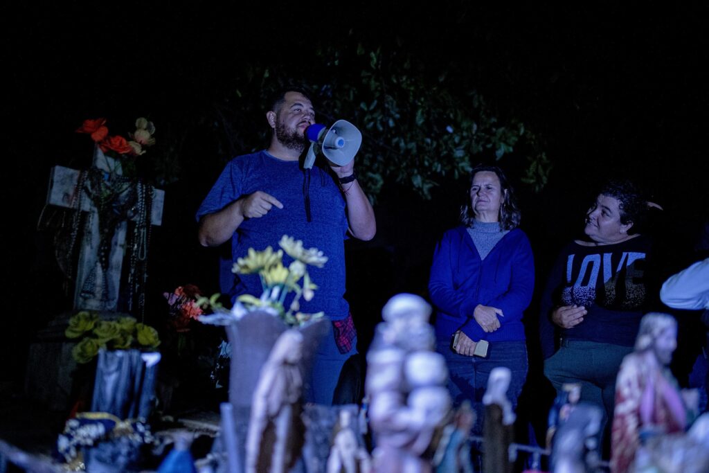 A imagem mostra uma cena noturna em um cemitério. Um homem com barba está falando em um megafone, apontando para a esquerda. Ele está vestindo uma camiseta azul e jeans. À sua direita, duas mulheres estão em pé. A primeira, vestindo um agasalho azul e uma blusa cinza, olha para a frente com uma expressão séria. A segunda mulher, usando uma blusa preta com a palavra "LOVE" escrita em letras brilhantes, olha para a primeira mulher. No fundo, à esquerda, há uma cruz decorada com flores e contas. O primeiro plano está escuro, mas sugere a presença de pequenas estátuas e arranjos florais, possivelmente em túmulos. A iluminação da cena é fraca, criando uma atmosfera sombria e misteriosa.