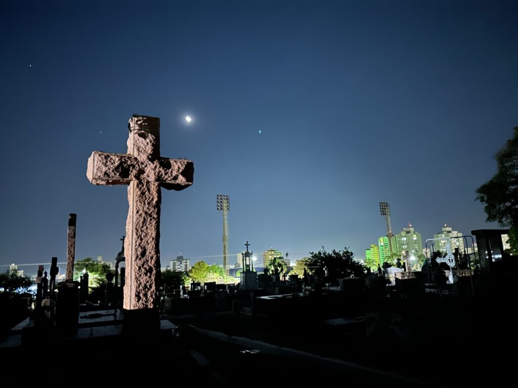 A imagem retrata um cemitério à noite, banhado pela luz da lua e de postes de iluminação distantes. Em primeiro plano, uma cruz de pedra imponente se destaca, com sua textura rústica e detalhes desgastados pelo tempo. Ao fundo, o horizonte da cidade se revela com prédios iluminados e torres de estádios, contrastando com a serenidade do local de descanso. O céu azul escuro, pontilhado por estrelas e a lua crescente, confere um ar melancólico e contemplativo à cena. A escuridão que envolve a maior parte do cemitério cria um jogo de luz e sombra, acentuando a atmosfera misteriosa e etérea do local.