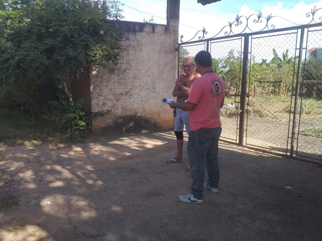 A imagem mostra duas pessoas em pé em um terreno de terra batida, com uma parede de alvenaria à esquerda e um portão de metal ornamentado à direita. O homem à esquerda, de óculos e calvo, está sem camisa e veste shorts. Ele cruza os braços e olha para o homem à sua frente. O homem à direita veste uma camiseta rosa com um logotipo circular nas costas e calças jeans. Ele segura um papel na mão e parece estar falando com o outro homem, possivelmente apresentando informações ou fazendo perguntas. O ambiente ao redor sugere uma área externa, possivelmente rural ou em uma zona menos urbanizada, com vegetação ao fundo e um céu azul com algumas nuvens. A luz do sol cria sombras distintas no chão.