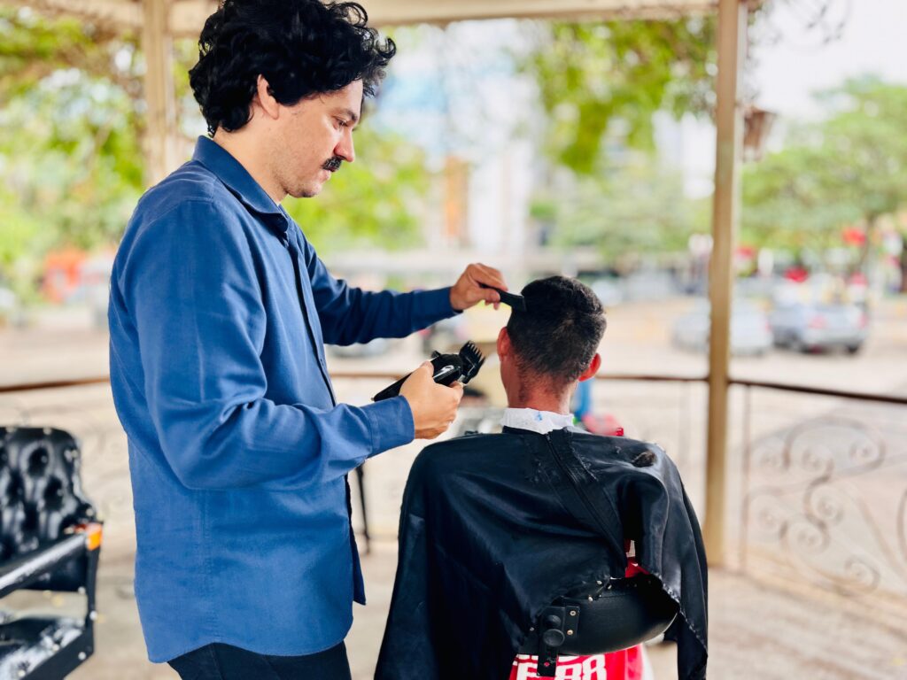 A imagem mostra um barbeiro cortando o cabelo de um cliente ao ar livre. O barbeiro, vestindo uma camisa azul e com um bigode característico, está concentrado em seu trabalho, segurando uma máquina de cortar cabelo em uma mão e um pente na outra. Ele está inclinando-se sobre o cliente, que está sentado de costas para a câmera, usando uma capa preta para proteger suas roupas. O ambiente sugere um local informal, possivelmente uma varanda ou um espaço aberto com cobertura, onde árvores e algumas construções podem ser vistas ao fundo, desfocadas. A luz natural ilumina a cena, criando um ambiente agradável e descontraído. O foco principal está na interação entre o barbeiro e o cliente, capturando o momento do corte de cabelo.
