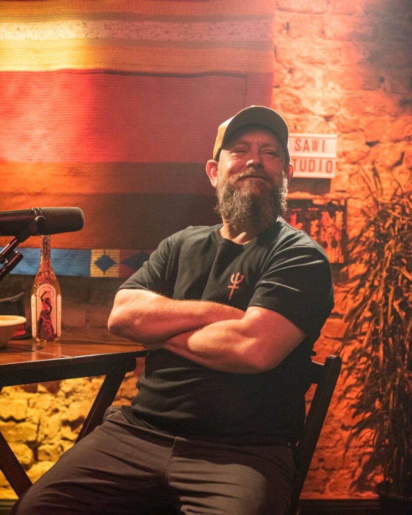 A imagem mostra um homem com barba e um boné, sentado em uma cadeira com os braços cruzados. Ele está vestindo uma camiseta preta com um símbolo vermelho no peito. Atrás dele, há uma parede de tijolos com uma placa que diz "SAWI STUDIO". Há também um microfone e uma garrafa em uma mesa. O fundo é decorado com um tecido colorido com listras horizontais em tons de laranja e amarelo. A iluminação da cena é quente e cria um ambiente acolhedor.