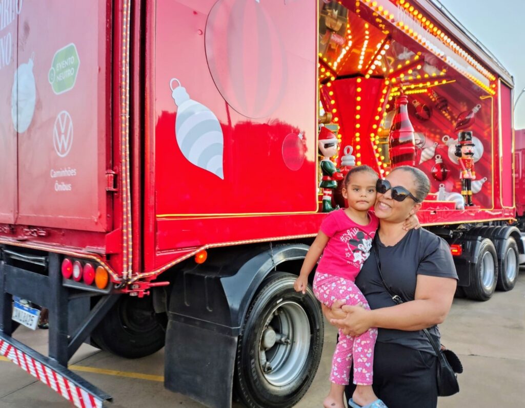 A imagem mostra uma mulher segurando uma menina em frente a um caminhão vermelho decorado com tema natalino. O caminhão possui luzes brilhantes, enfeites natalinos como bolas e bonecos de quebra-nozes, e uma garrafa icônica da Coca-Cola. Na lateral do caminhão, há o logo da Volkswagen com os dizeres "Caminhões Ônibus" e um selo "Evento Neutro". A menina veste uma camiseta rosa com estampa do Mickey Mouse e calças cor-de-rosa com corações. A mulher usa óculos escuros e um sorriso no rosto, abraçando a menina carinhosamente. O fundo sugere um ambiente externo, possivelmente um evento ou parada natalina.