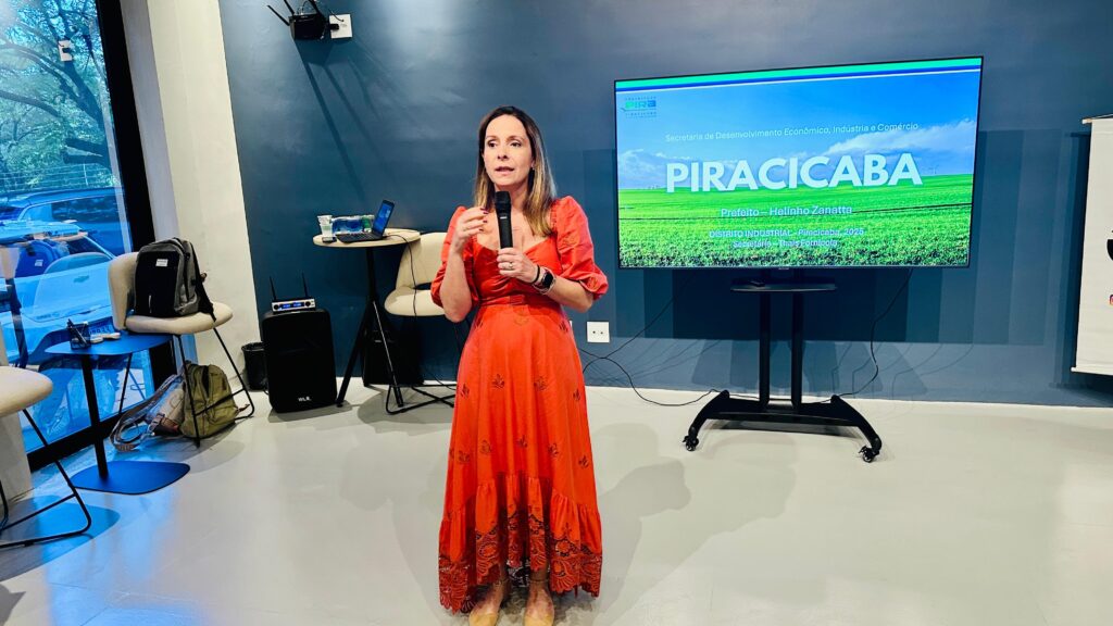A imagem mostra uma mulher palestrante em um evento, falando em um microfone enquanto está em um palco ou área de apresentação. Ela veste um vestido laranja com detalhes em azul e vermelho, e parece estar explicando ou apresentando algo para o público. Atrás dela, há uma grande tela que exibe um slide de apresentação. No slide, é possível ler informações relacionadas à Secretaria de Desenvolvimento Econômico, Indústria e Comércio de Piracicaba, com menção ao prefeito Helinho Zanatta e à secretária Thais Fornícola. Também aparece o título "Distrito Industrial - Piracicaba, 2025". O ambiente parece ser uma sala de conferências ou auditório, com equipamentos de som e cadeiras visíveis. A iluminação é clara e destaca a palestrante e a tela. A atmosfera sugere uma reunião ou evento oficial voltado para o desenvolvimento econômico local.