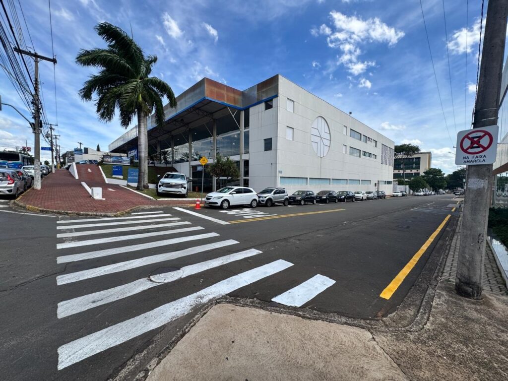 A imagem mostra uma rua com uma faixa de pedestres em primeiro plano. Ao lado da rua, há um prédio moderno com janelas amplas e uma entrada coberta. Uma palmeira alta se destaca em frente ao prédio. Vários carros estão estacionados na rua e na entrada do prédio. No canto superior direito, há uma placa de trânsito com um símbolo de proibição e a inscrição "NA FAIXA AMARELA". O céu está azul com algumas nuvens.