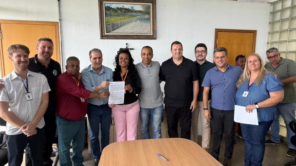 Na imagem, um grupo de doze pessoas posa em um ambiente de escritório, transmitindo a ideia de uma ocasião formal ou comemorativa. Algumas delas, posicionadas ao centro, seguram um documento, sugerindo a assinatura de um acordo ou a conclusão de uma reunião importante. Os participantes estão vestidos de forma variada, entre trajes casuais e sociais, e alguns usam crachás de identificação. O espaço é simples e organizado, com paredes brancas, portas de madeira, uma mesa em primeiro plano e um quadro de paisagem pendurado na parede. A cena reflete colaboração e conquista coletiva em um contexto profissional.