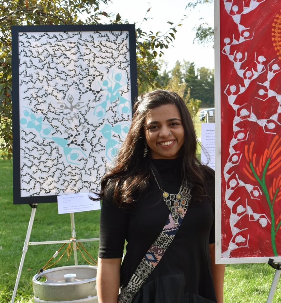 A mulher de pele morena, com cabelos longos e escuros, está sorrindo para a câmera. Ela usa um vestido preto de gola alta e uma echarpe colorida com padrões geométricos que cruza o peito. Um colar com pedras de diversas cores e formatos complementa seu visual. À sua esquerda, há um quadro grande com uma arte abstrata em tons de preto, branco e azul claro, com um padrão que lembra galhos e figuras estilizadas. O quadro está apoiado em um tripé. À sua direita, outro quadro exibe uma pintura vibrante em fundo vermelho, com figuras brancas estilizadas que parecem dançar em círculo, lembrando um estilo de arte popular. O cenário é ao ar livre, com gramado verde, árvores ao fundo e um céu parcialmente nublado. Há também um barril metálico visível no canto inferior esquerdo.