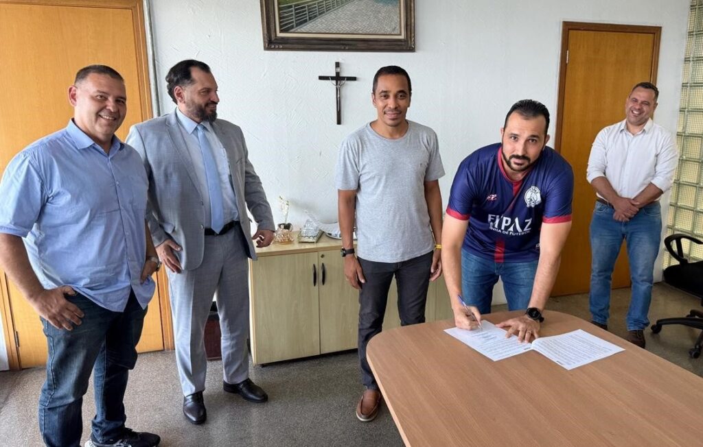 A imagem mostra cinco homens em um escritório, com um homem de camisa azul e jeans, um homem de terno cinza e gravata azul clara, um homem de camiseta cinza e jeans escuros, um homem de camisa de time de futebol azul e vermelha e jeans, e um homem de camisa branca e jeans. O homem de camisa de time está assinando um documento em uma mesa de madeira. Há um crucifixo na parede atrás dos homens, e um quadro com uma imagem de uma passarela com árvores. O ambiente parece ser um escritório formal, com armários e uma porta de madeira.