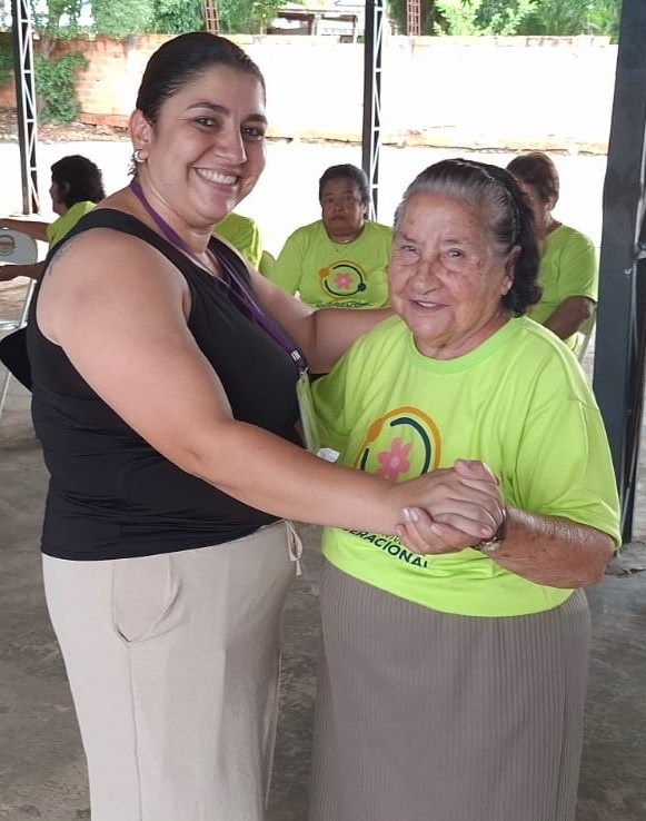 A imagem mostra duas mulheres, uma mais jovem e outra mais idosa, dançando juntas em um ambiente externo com cobertura. A mulher mais jovem, à esquerda, veste uma blusa preta sem mangas e calças bege. Ela tem cabelos escuros presos e está sorrindo para a câmera, com o braço estendido para segurar a mão da outra mulher. A mulher mais idosa, à direita, veste uma camiseta verde clara com um logotipo que parece ser uma flor dentro de um círculo. Ela tem cabelos grisalhos presos para trás e um sorriso gentil. Ela está segurando a mão da mulher mais jovem e parece estar aproveitando a dança. Ao fundo, outras pessoas, também vestindo camisetas verdes semelhantes, estão sentadas e observando. O ambiente sugere um evento comunitário ou uma atividade recreativa para idosos.