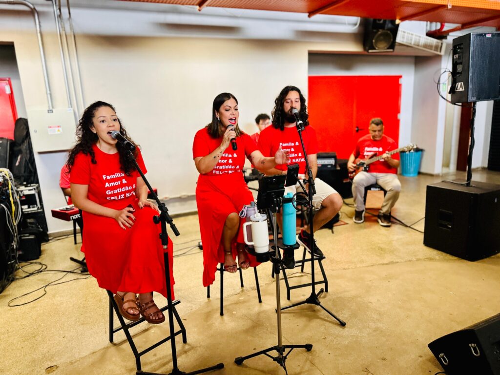 Na imagem, um grupo de pessoas está se apresentando musicalmente, possivelmente em um evento ou ensaio. Três pessoas estão em destaque, sentadas em cadeiras altas e vestindo camisetas vermelhas com a mesma estampa: "Família Amor & Gratidão & Feliz Natal". A pessoa mais à esquerda é uma mulher com cabelos cacheados escuros, cantando em um microfone. Ao seu lado, uma mulher com cabelos castanhos escuros, também cantando em um microfone. O terceiro em destaque é um homem com cabelos longos e barba, que também está cantando em um microfone. No fundo, um homem toca guitarra, e ao lado dele, outro homem, possivelmente um músico de apoio, está visível. A cena parece ser em um ambiente interno com piso de concreto e paredes claras, com elementos de palco como caixas de som e equipamentos de áudio. Há também garrafas de água em um suporte próximo aos cantores.