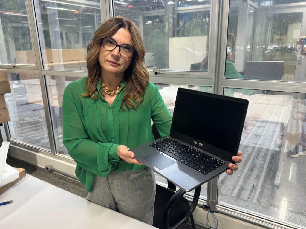 A mulher está segurando um laptop preto com a marca "DATEN" visível. Ela está usando uma blusa verde transparente com gola alta dourada e calças xadrez cinza. Ela usa óculos pretos e tem cabelo castanho ondulado. Ela está em um ambiente interno com grandes janelas ao fundo, que mostram uma área externa com algumas caixas empilhadas e reflexos de carros.
