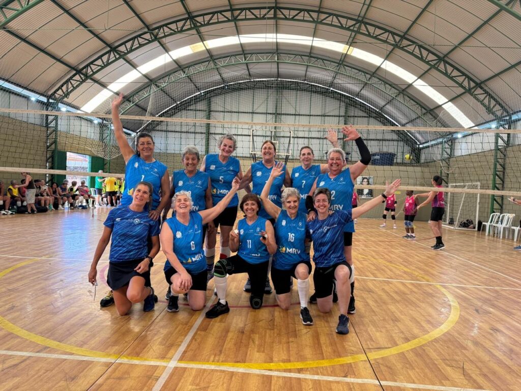 A imagem mostra um time de vôlei feminino posando para uma foto em uma quadra interna. As jogadoras, todas mulheres mais velhas, vestem uniformes azuis com o número de cada uma visível. Algumas estão em pé e outras ajoelhadas na frente, todas sorrindo e acenando para a câmera. O fundo exibe a quadra de vôlei com uma rede esticada e a estrutura do telhado acima. Ao fundo, outras pessoas podem ser vistas sentadas nas arquibancadas e algumas jogadoras em quadras adjacentes. A imagem transmite uma atmosfera de camaradagem e esporte.