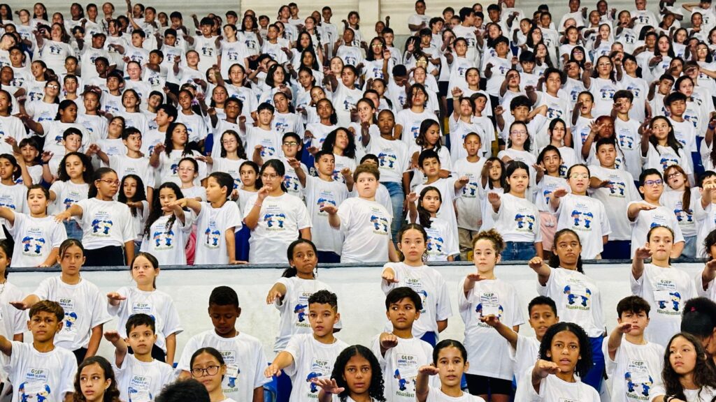 A imagem mostra um grande grupo de crianças e adolescentes em pé, em várias fileiras, todos vestindo camisetas brancas com o mesmo design. O design da camiseta parece incluir um logotipo com as letras "GCEP" e a inscrição "PELOTÃO ESCOLAR" e "GUARDA CIVIL". As crianças estão em um espaço interno, possivelmente uma quadra ou ginásio, com uma parede ao fundo. A maioria das crianças está com a mão direita levantada e estendida para a frente, com a palma virada para baixo, em um gesto que pode ser de saudação, juramento ou algum tipo de sinal combinado. Elas parecem estar em um evento organizado, talvez uma cerimônia escolar ou uma atividade cívica. A iluminação é uniforme, sugerindo um ambiente interno bem iluminado. A expressão facial das crianças varia, mas muitas parecem atentas e engajadas.