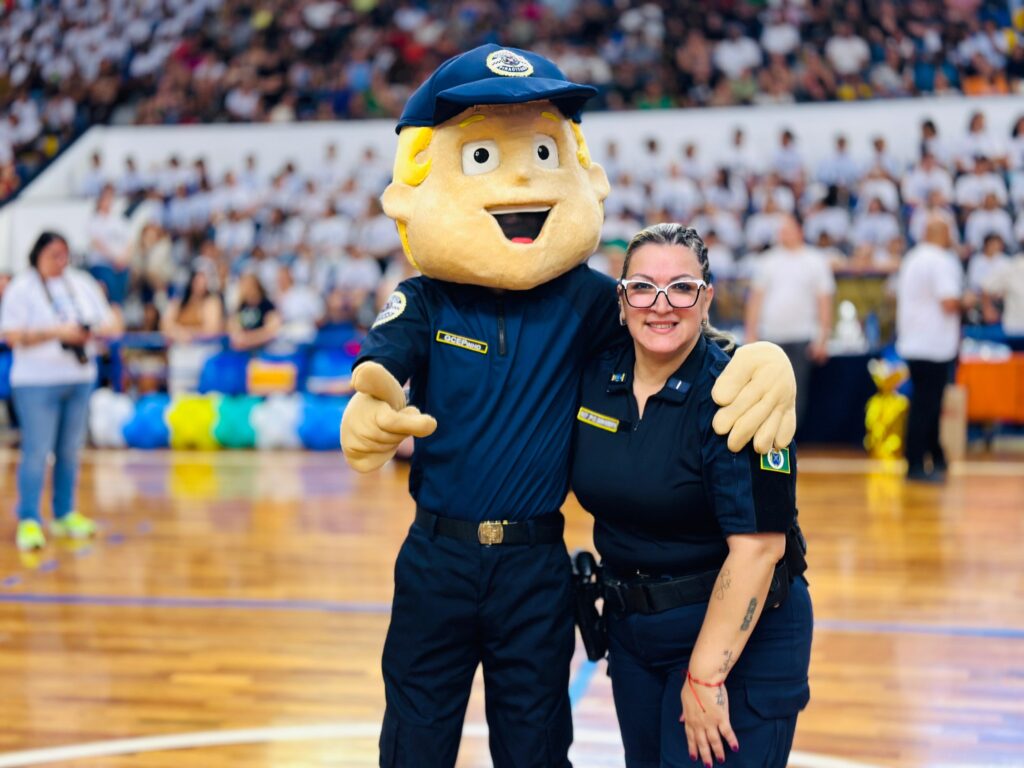 A imagem mostra um mascote com uniforme de policial e uma policial sorrindo para a câmera. O mascote, com um chapéu azul e um macacão escuro, está com a mão no ombro da policial. A policial, usando óculos grandes e um uniforme semelhante, também sorri. Ambos estão em uma quadra de esportes, com uma plateia desfocada ao fundo. Há balões coloridos dispostos no chão atrás deles.