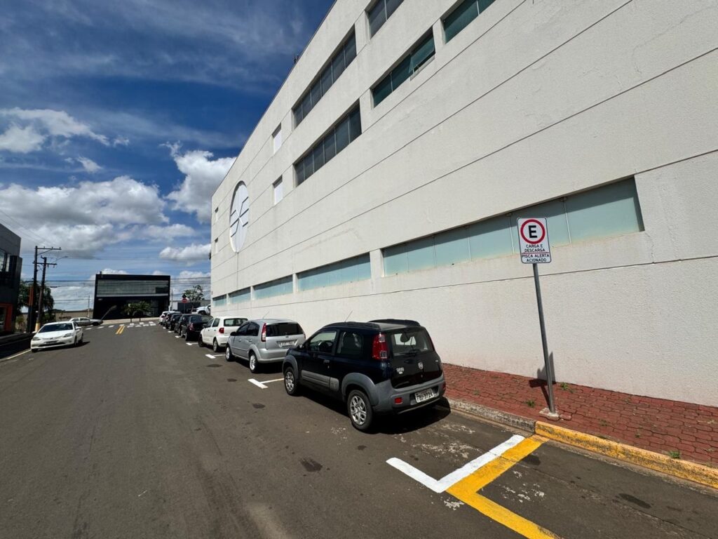 A imagem mostra uma rua com vários carros estacionados ao lado de um prédio branco moderno. O céu está azul com algumas nuvens brancas. Há uma placa de trânsito com um "E" dentro de um círculo vermelho, indicando "Carga e Descarga" e "Pisca Alerta Ligado". A rua é asfaltada e há uma faixa amarela e branca pintada no chão, indicando a área de estacionamento. No fundo, vê-se outro prédio escuro e uma linha de postes de eletricidade.
